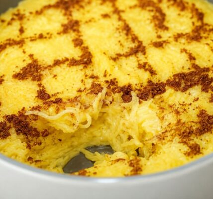 Uma travessa redonda cinza contendo aletria cremosa amarela, uma sobremesa tradicional portuguesa feita com fios finos de massa, polvilhada com canela em pó desenhada em ziguezague, com uma porção retirada no centro revelando a textura.