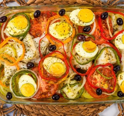 Uma travessa de vidro transparente exibe uma porção generosa de bacalhau à Gomes de Sá. O prato, visto de cima, revela lascas de bacalhau misturadas com batatas, rodelas de cebola e tomates. Anéis coloridos de pimentão vermelho, amarelo e verde criam padrões geométricos. Metades de ovos cozidos com gemas amarelas e azeitonas pretas estão espalhadas sobre a superfície. A travessa repousa sobre um jogo americano trançado de vime. Ao fundo, um guardanapo de linho azul com listras brancas e um pedaço de tecido denim. A iluminação é natural e brilhante.
