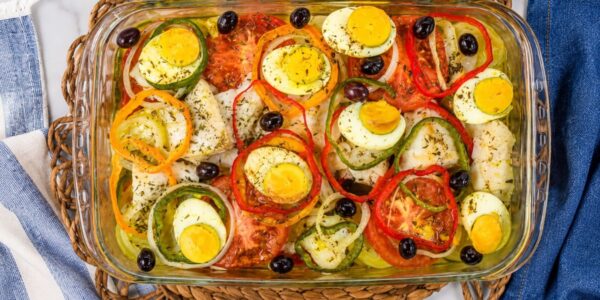 Uma travessa de vidro transparente exibe uma porção generosa de bacalhau à Gomes de Sá. O prato, visto de cima, revela lascas de bacalhau misturadas com batatas, rodelas de cebola e tomates. Anéis coloridos de pimentão vermelho, amarelo e verde criam padrões geométricos. Metades de ovos cozidos com gemas amarelas e azeitonas pretas estão espalhadas sobre a superfície. A travessa repousa sobre um jogo americano trançado de vime. Ao fundo, um guardanapo de linho azul com listras brancas e um pedaço de tecido denim. A iluminação é natural e brilhante.