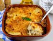 Uma travessa de barro cheia de bacalhau com natas gratinado, com uma crosta dourada e queijo derretido, com uma colher de servir retirando uma porção do molho cremoso. Ao fundo, um pano xadrez azul, talheres e azeite.