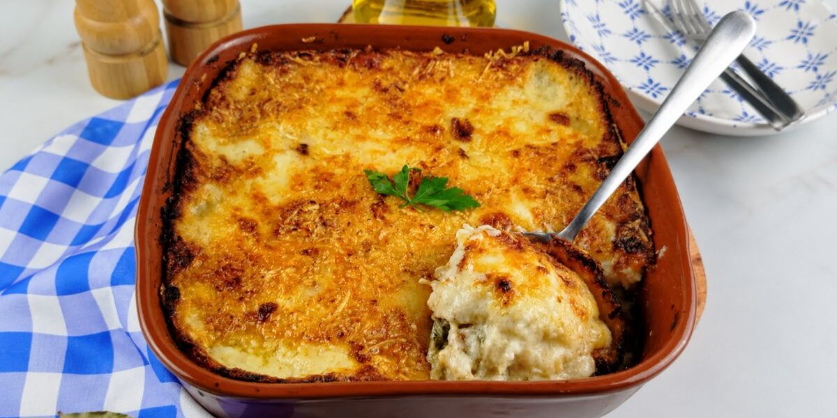 Uma travessa de barro cheia de bacalhau com natas gratinado, com uma crosta dourada e queijo derretido, com uma colher de servir retirando uma porção do molho cremoso. Ao fundo, um pano xadrez azul, talheres e azeite.