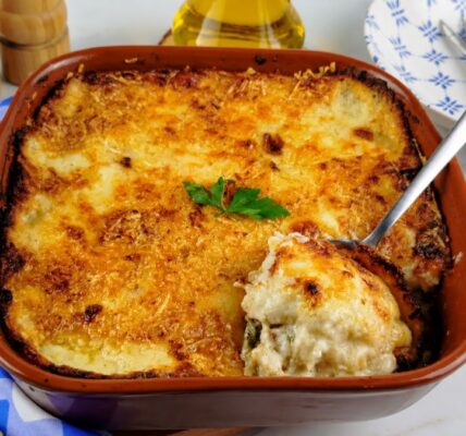 Uma travessa de barro cheia de bacalhau com natas gratinado, com uma crosta dourada e queijo derretido, com uma colher de servir retirando uma porção do molho cremoso. Ao fundo, um pano xadrez azul, talheres e azeite.