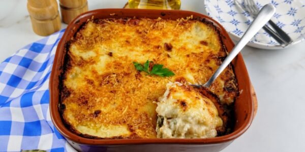 Uma travessa de barro cheia de bacalhau com natas gratinado, com uma crosta dourada e queijo derretido, com uma colher de servir retirando uma porção do molho cremoso. Ao fundo, um pano xadrez azul, talheres e azeite.
