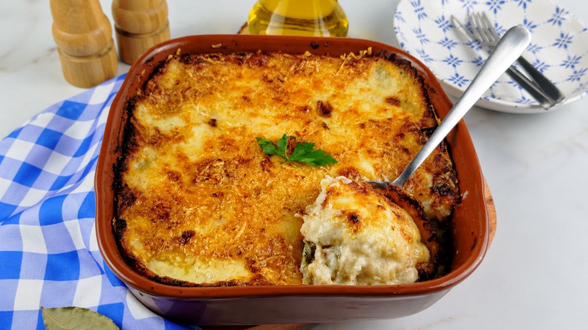 Uma travessa de barro cheia de bacalhau com natas gratinado, com uma crosta dourada e queijo derretido, com uma colher de servir retirando uma porção do molho cremoso. Ao fundo, um pano xadrez azul, talheres e azeite.