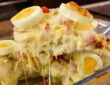 Travessa de vidro contendo Batata à Portuguesa gratinada, com camadas visíveis de molho branco cremoso, presunto e queijo mussarela derretido, decorada com rodelas de ovos cozidos e azeitonas verdes. Uma porção está sendo levantada por uma espátula, revelando o queijo esticando.
