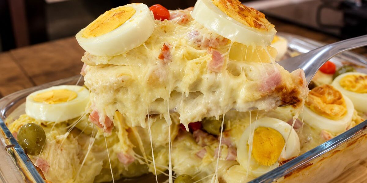 Travessa de vidro contendo Batata à Portuguesa gratinada, com camadas visíveis de molho branco cremoso, presunto e queijo mussarela derretido, decorada com rodelas de ovos cozidos e azeitonas verdes. Uma porção está sendo levantada por uma espátula, revelando o queijo esticando.