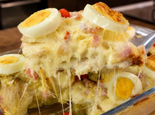 Travessa de vidro contendo Batata à Portuguesa gratinada, com camadas visíveis de molho branco cremoso, presunto e queijo mussarela derretido, decorada com rodelas de ovos cozidos e azeitonas verdes. Uma porção está sendo levantada por uma espátula, revelando o queijo esticando.