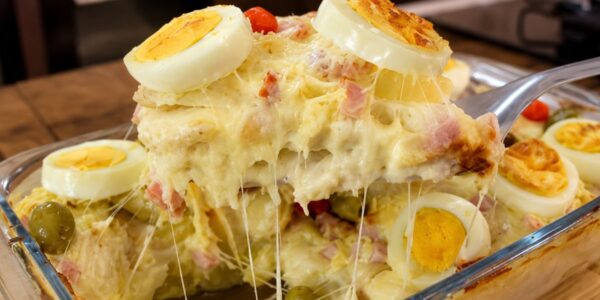 Travessa de vidro contendo Batata à Portuguesa gratinada, com camadas visíveis de molho branco cremoso, presunto e queijo mussarela derretido, decorada com rodelas de ovos cozidos e azeitonas verdes. Uma porção está sendo levantada por uma espátula, revelando o queijo esticando.