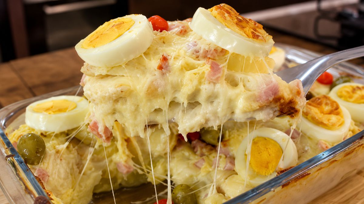 Travessa de vidro contendo Batata à Portuguesa gratinada, com camadas visíveis de molho branco cremoso, presunto e queijo mussarela derretido, decorada com rodelas de ovos cozidos e azeitonas verdes. Uma porção está sendo levantada por uma espátula, revelando o queijo esticando.