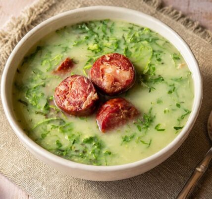Uma tigela de cerâmica rústica cheia de caldo verde tradicional português sobre um pano de linho. O caldo é um creme de batata espesso de cor verde pálida misturado com tiras finas de couve verde escura. No topo da sopa estão três rodelas grossas e grelhadas de linguiça portuguesa defumada. Ao lado da tigela há uma colher de metal prateada e fatias de pão rústico em uma tábua de madeira ao fundo.