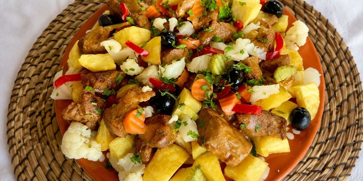 Prato tradicional de carne de porco à portuguesa servido em travessa de barro com cubos de batata assada, pickles variados, azeitonas pretas e coentros picados.