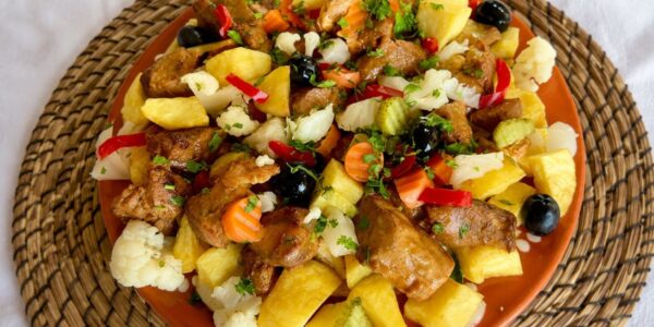 Prato tradicional de carne de porco à portuguesa servido em travessa de barro com cubos de batata assada, pickles variados, azeitonas pretas e coentros picados.