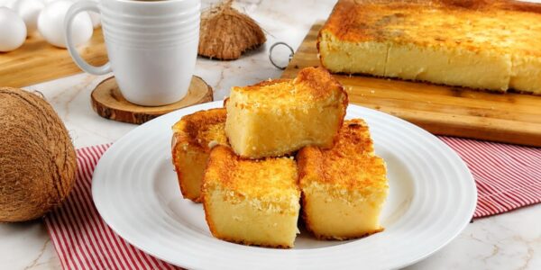 Fatias quadradas de bolo de coco com cobertura tostada em prato branco, café ao fundo.