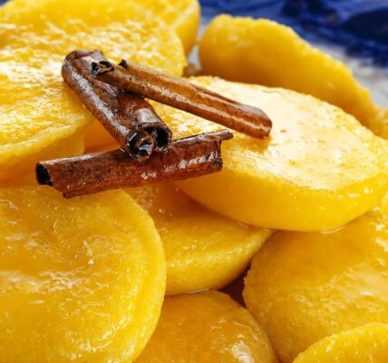 Close em doces papos de anjo amarelos brilhantes imersos em calda transparente com canela em pau por cima sobre prato azul.