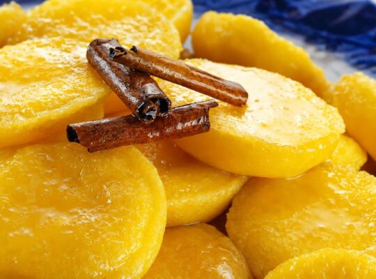 Close em doces papos de anjo amarelos brilhantes imersos em calda transparente com canela em pau por cima sobre prato azul.
