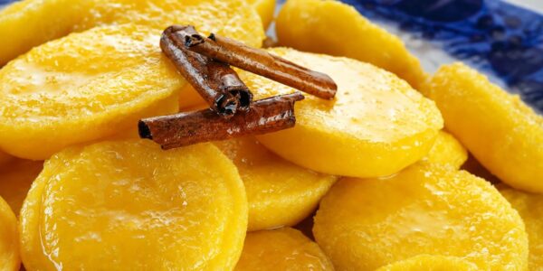 Close em doces papos de anjo amarelos brilhantes imersos em calda transparente com canela em pau por cima sobre prato azul.