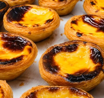 Vários pastéis de nata artesanais dispostos sobre papel manteiga, exibindo casca de massa folhada dourada e crocante com recheio de creme de ovos caramelizado no topo, ideais para sobremesa.