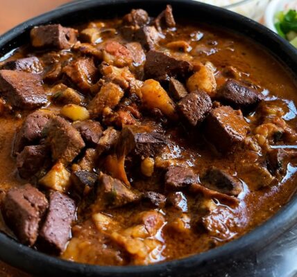 Sarapatel de porco servido em uma panela de barro preta com molho encorpado e miúdos picados. Ao fundo, acompanhamentos como farofa crocante e vinagrete fresco em tigelas brancas.