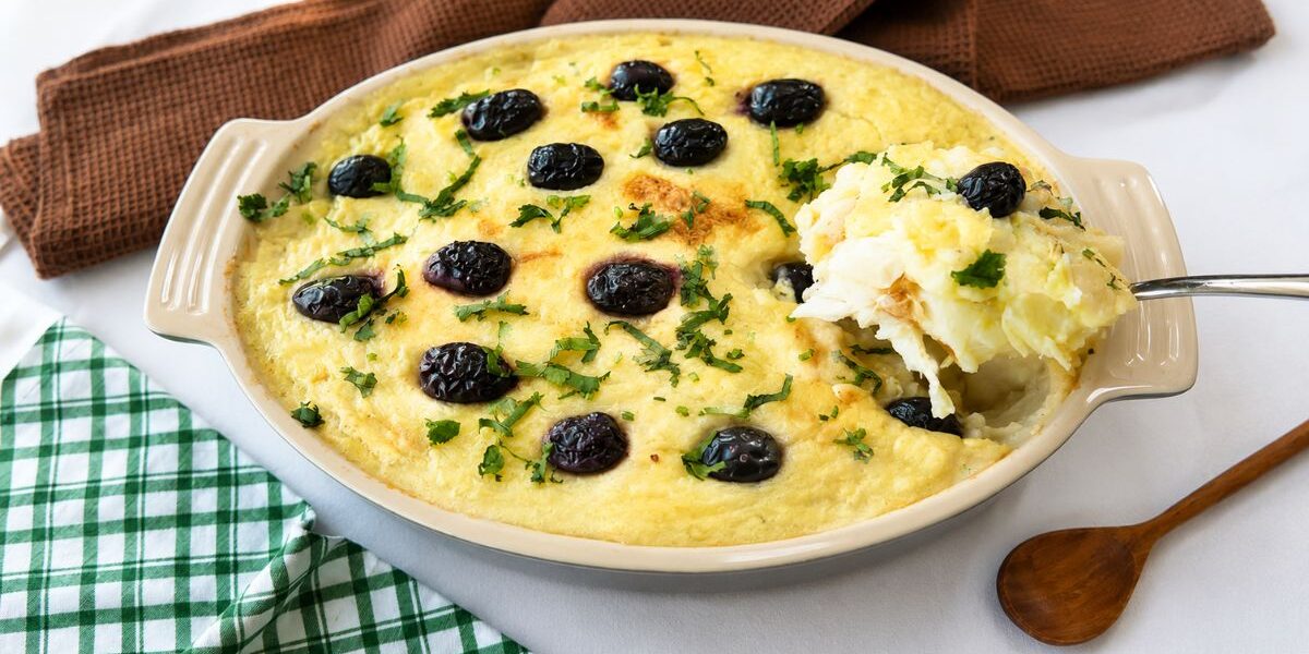 Uma travessa refratária oval de cerâmica contendo Bacalhau à Zé do Pipo gratinado. O prato é coberto com um purê de batata dourado, azeitonas pretas inteiras e salsinha picada. Uma colher de metal retira uma porção cremosa do canto direito. Ao fundo, um guardanapo de linho marrom e um pano xadrez verde e branco sobre uma superfície branca. Uma colher de madeira repousa à direita.