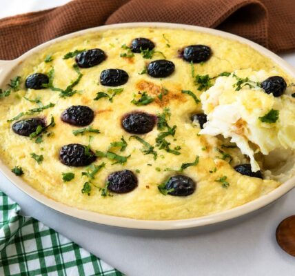 Uma travessa refratária oval de cerâmica contendo Bacalhau à Zé do Pipo gratinado. O prato é coberto com um purê de batata dourado, azeitonas pretas inteiras e salsinha picada. Uma colher de metal retira uma porção cremosa do canto direito. Ao fundo, um guardanapo de linho marrom e um pano xadrez verde e branco sobre uma superfície branca. Uma colher de madeira repousa à direita.