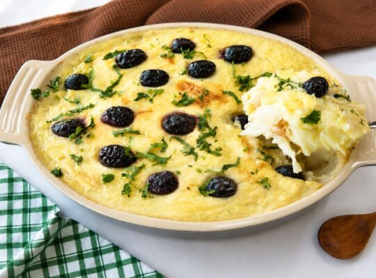 Uma travessa refratária oval de cerâmica contendo Bacalhau à Zé do Pipo gratinado. O prato é coberto com um purê de batata dourado, azeitonas pretas inteiras e salsinha picada. Uma colher de metal retira uma porção cremosa do canto direito. Ao fundo, um guardanapo de linho marrom e um pano xadrez verde e branco sobre uma superfície branca. Uma colher de madeira repousa à direita.