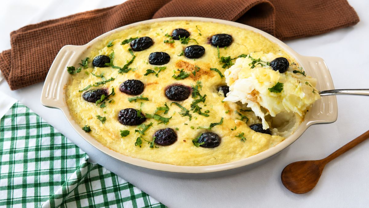 Uma travessa refratária oval de cerâmica contendo Bacalhau à Zé do Pipo gratinado. O prato é coberto com um purê de batata dourado, azeitonas pretas inteiras e salsinha picada. Uma colher de metal retira uma porção cremosa do canto direito. Ao fundo, um guardanapo de linho marrom e um pano xadrez verde e branco sobre uma superfície branca. Uma colher de madeira repousa à direita.