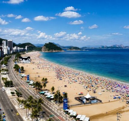 Fotografia panorâmica do alto da praia de Copacabana repleta de banhistas, mostrando a avenida Atlântica, calçadão, quiosques, mar azul e morros ao fundo sob céu ensolarado.