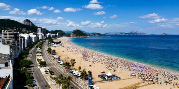 Fotografia panorâmica do alto da praia de Copacabana repleta de banhistas, mostrando a avenida Atlântica, calçadão, quiosques, mar azul e morros ao fundo sob céu ensolarado.