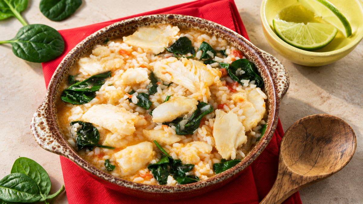 Uma tigela de cerâmica rústica cheia de arroz de bacalhau cremoso com lascas de bacalhau espessas e folhas de espinafre, colocada sobre um guardanapo vermelho numa mesa de pedra clara, com lima e colher de madeira ao lado.