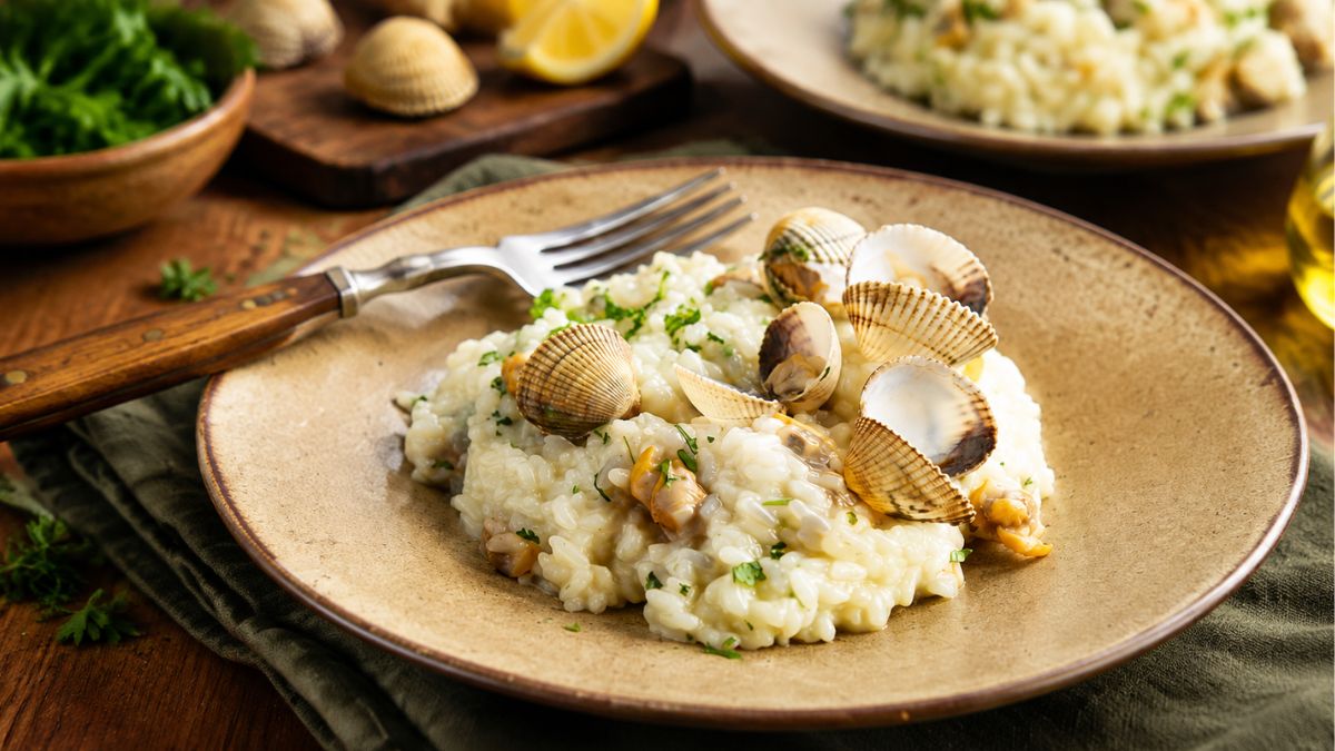 Prato rústico com arroz de berbigão e camarão, guarnecido com cascas e coentros picados, acompanhado por um garfo de madeira.