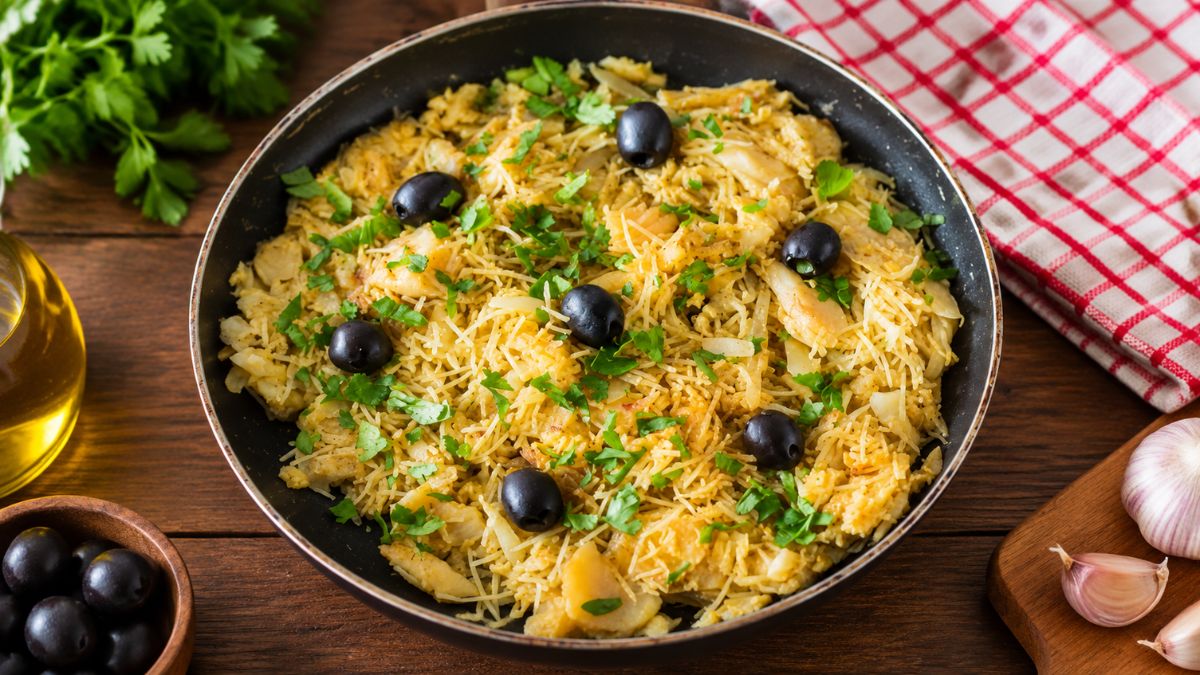 Frigideira preta rústica com uma porção de Bacalhau à Brás cremoso, misturado com batata palha, ovo e azeitonas pretas, polvilhado com salsa fresca. O fundo é de madeira com alho, azeite e um pano xadrez.