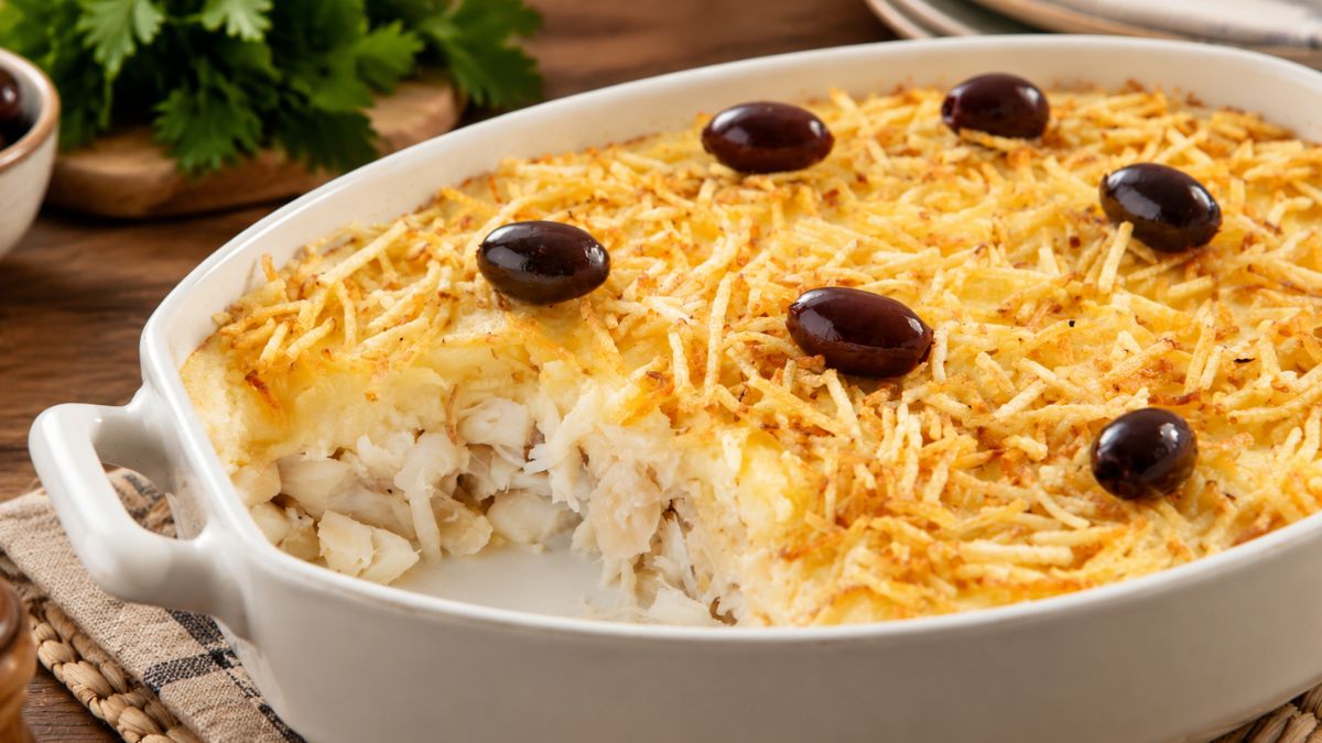 Bacalhau gratinado numa travessa branca refratária com camada cremosa de natas, cobertura dourada de batata palha estaladiça e decorado com azeitonas pretas inteiras.