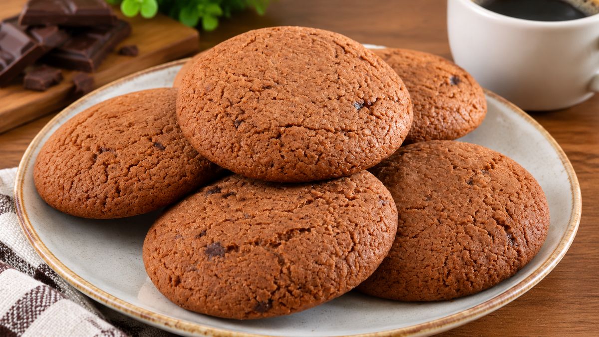 Prato raso de cerâmica com bolachas de canela caseiras redondas e douradas, acompanhado por uma chávena de café e pedaços de chocolate ao fundo.