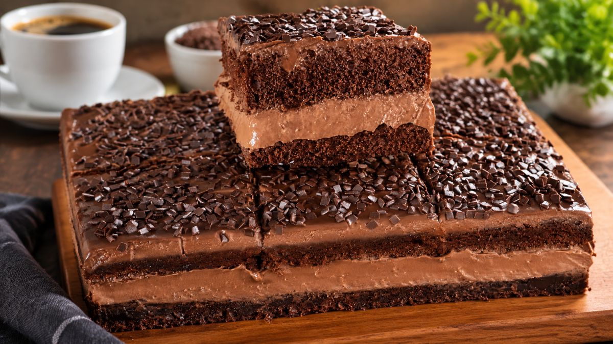 Bolo de chocolate retangular com recheio cremoso e cobertura de granulado de chocolate, disposto numa tábua de madeira com uma chávena de café ao fundo.
