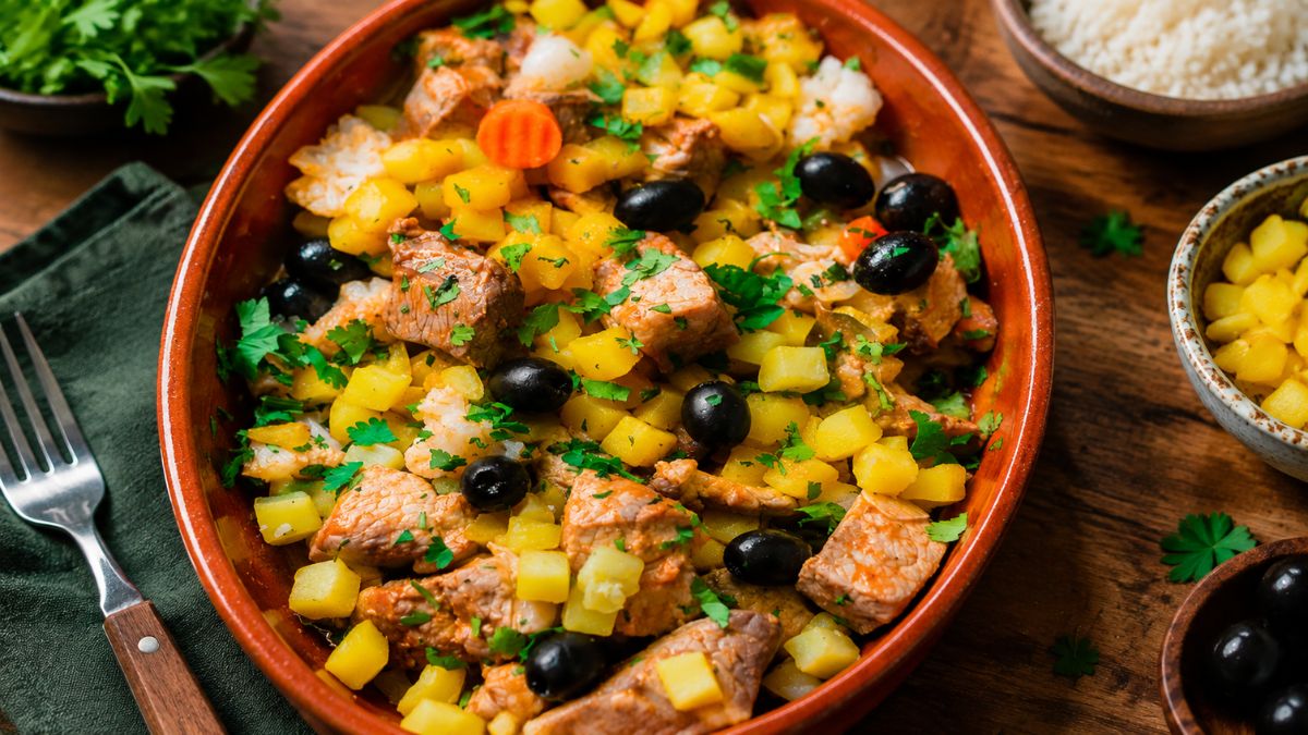 Travessa de barro com carne de porco à portuguesa enfeitada com batatas em cubos, azeitonas pretas e coentros frescos picados.