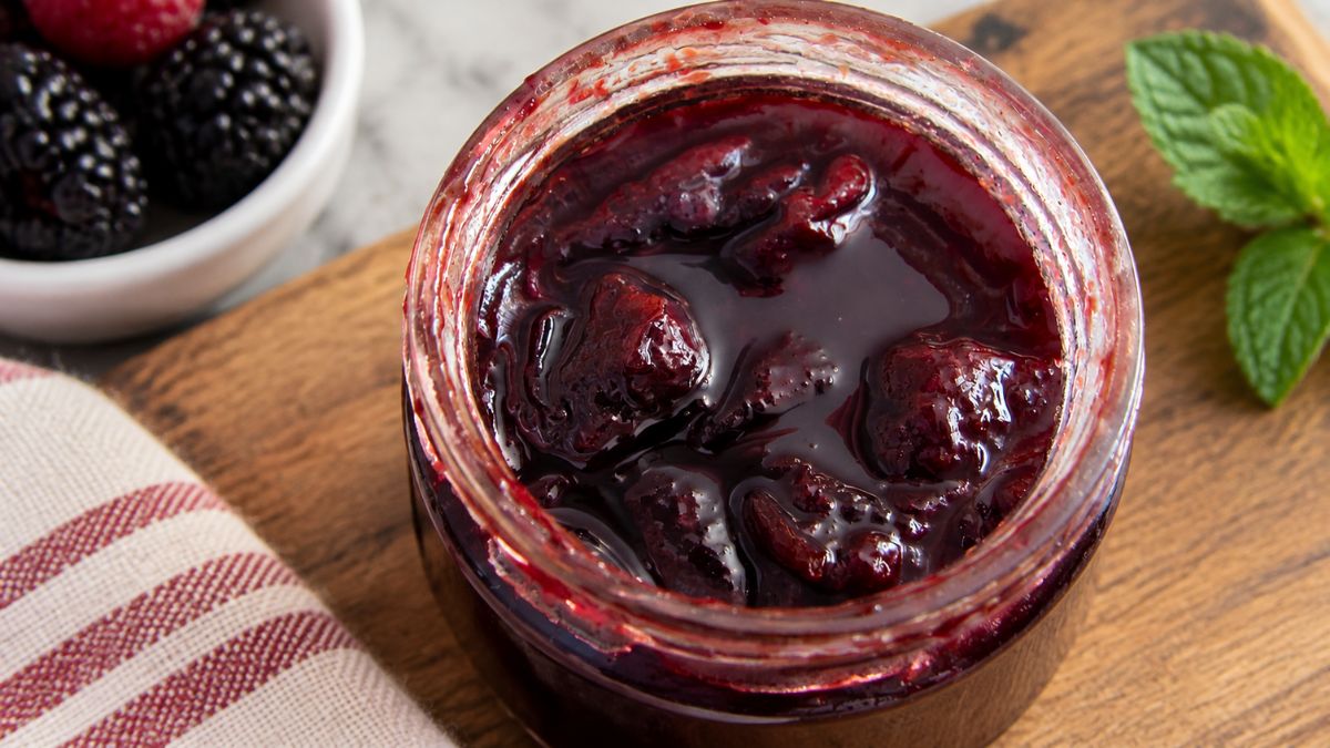 Vista superior de um frasco de vidro aberto com doce de ameixa caseiro e pedaços visíveis de fruta, colocado sobre uma tábua de madeira com mirtilos e hortelã ao fundo.