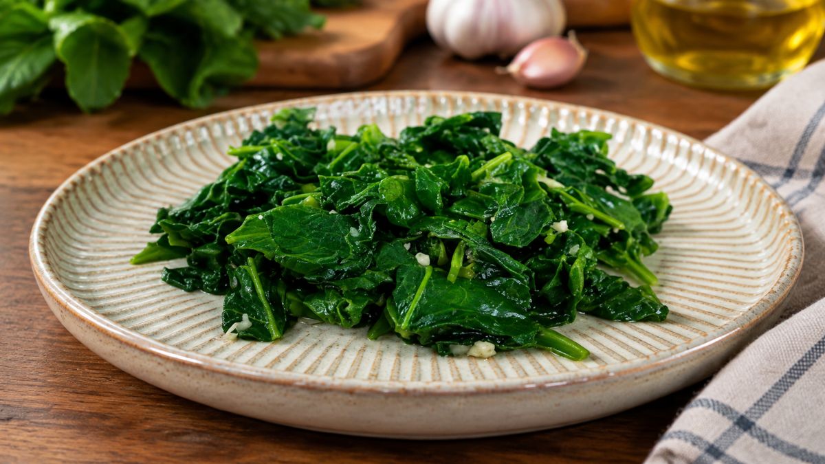 Um prato de cerâmica artesanal, com bordas listradas, apresenta uma porção generosa de espinafres salteados, de cor verde vibrante, com pedaços visíveis de alho picado. Ao fundo, uma mesa de madeira rústica mostra espinafres frescos, alho e uma garrafa de azeite.