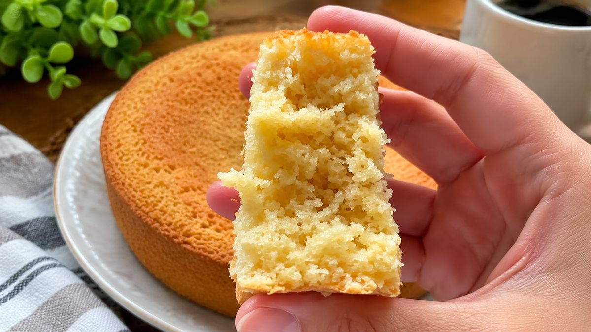close em uma mão feminina segurando uma fatia generosa de bolo de aveia mostrando a textura interna aerada e macia. Ao fundo está o bolo de aveia inteiro, um pano de prato xadrez, uma xícara branca e vegetação, sobre uma superfície de madeira.