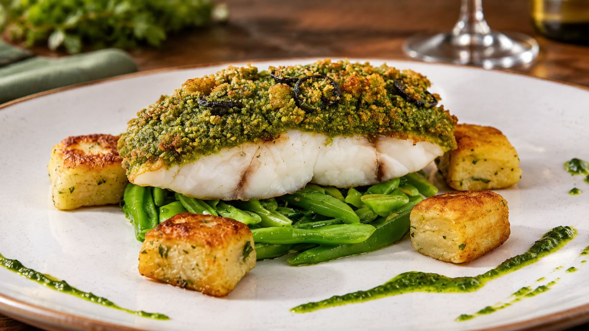 Filé de peixe branco suculento coberto por uma apetitosa crosta verde de ervas e alcaparras, servido sobre uma cama de vagem salteada e acompanhado de cubos de batatas douradas e crocantes, tudo disposto num prato de cerâmica branco com detalhes em marrom, focado para destacar a textura e os ingredientes frescos.