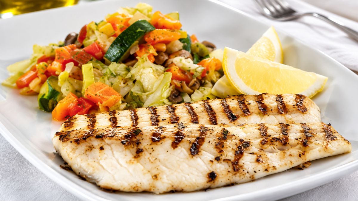 Close-up em um prato branco de filetes de peixe branco perfeitamente grelhados com marcas de grelha douradas, servidos com uma colorida salada de vegetais picados refogados com abobrinha, cenoura, pimentão e cogumelos, acompanhados por dois fatias de limão fresco e talheres no fundo desfocado em uma mesa rústica.