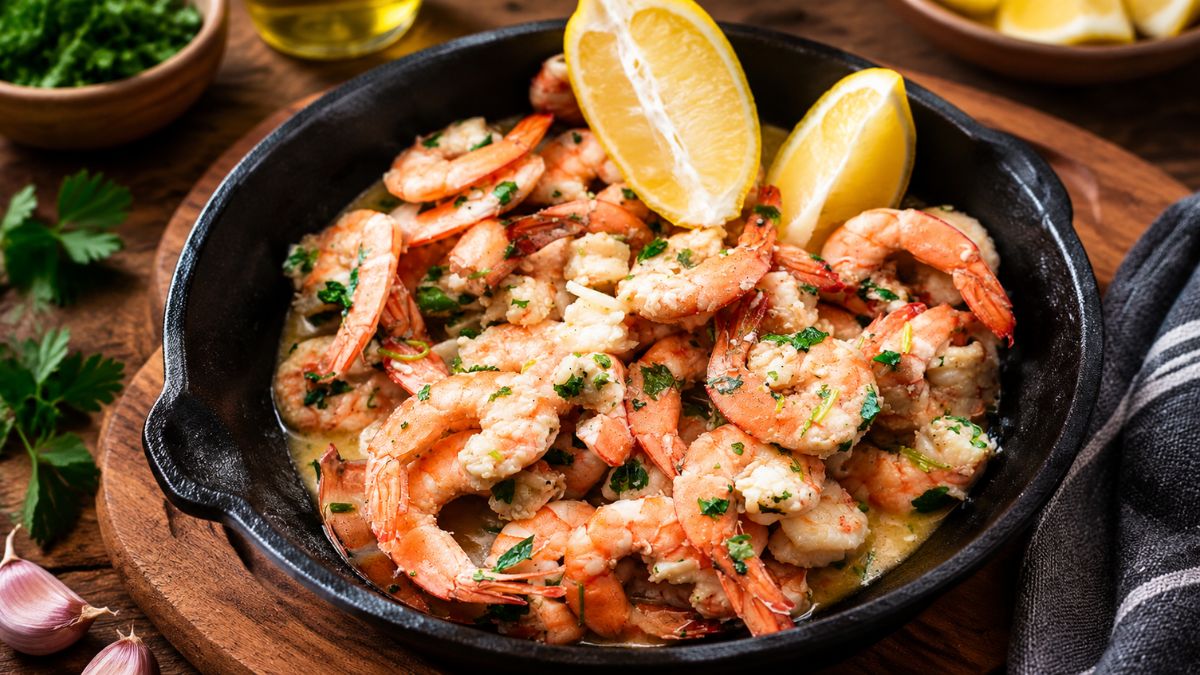 Uma frigideira de ferro preta cheia de camarões médios fritos com alho, regados com azeite e salpicados com salsa fresca picada. Duas fatias de limão siciliano estão no topo. A frigideira está sobre uma tábua de madeira redonda, com dentes de alho e um guardanapo de pano desfocado ao fundo.