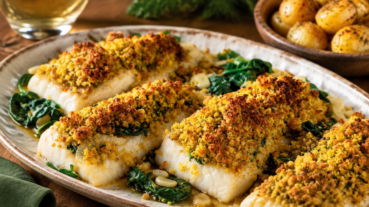 Lombos de bacalhau suculentos assados no forno com uma crosta crocante e dourada de pão de abóbora e nozes sobre uma cama de espinafres frescos salteados com alho. O prato está servido numa travessa rústica de cerâmica acompanhado por batatas primor assadas com ervas.