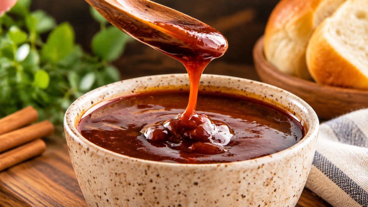 Taça de cerâmica com molho barbecue caseiro denso e brilhante a cair de uma colher de madeira sobre mesa rústica.
