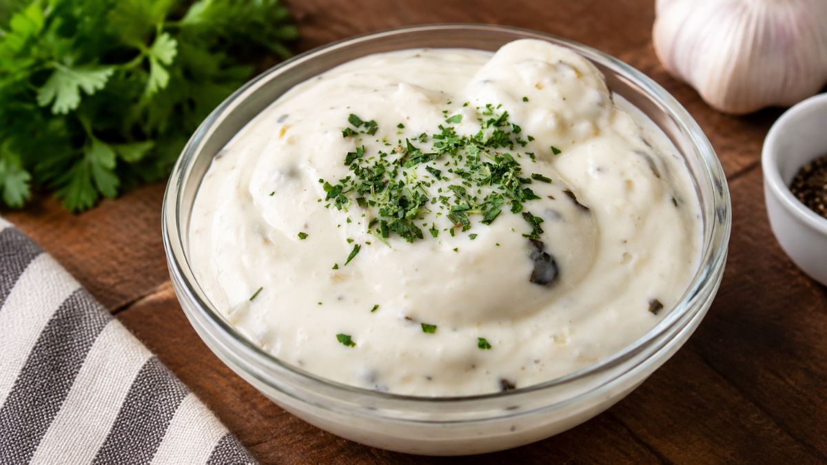 Tigela de vidro com molho de alho cremoso polvilhado com salsa picada, acompanhada por dentes de alho ao fundo.