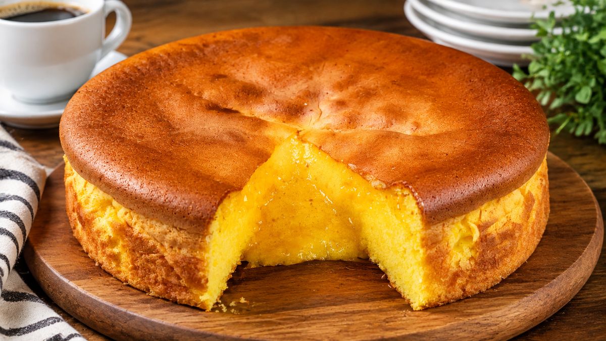Fotografia em close-up de um autêntico Pão de Ló de Ovar tradicional português, servido numa tábua de madeira redonda. O bolo tem uma côdea dourada e está cortado, revelando o seu interior extremamente húmido, cremoso e amarelo intenso, conhecido como pito. Ao fundo, vê-se uma chávena de café branco e pratos empilhados.