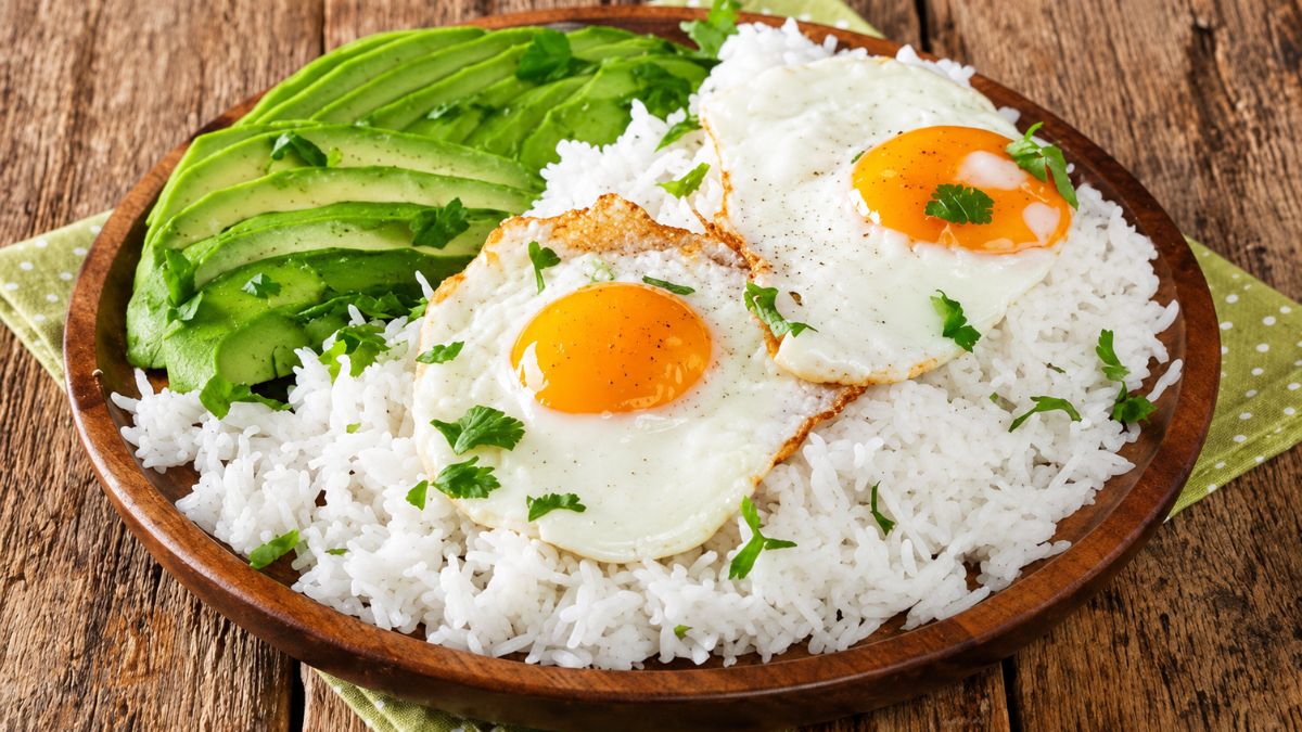 Prato rústico de madeira contendo uma base de arroz branco solto, coberto com dois ovos estrelados com gema mole e fatias de abacate fresco fatiado. O prato está polvilhado com coentros frescos picados sobre uma mesa de madeira.