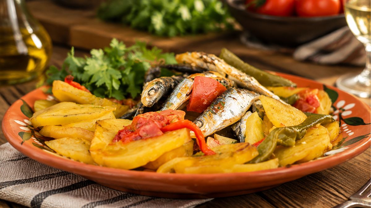 Sardinhas assadas portuguesas com batatas douradas, pimentos e coentros frescos servidas num prato de barro tradicional sobre mesa de madeira.