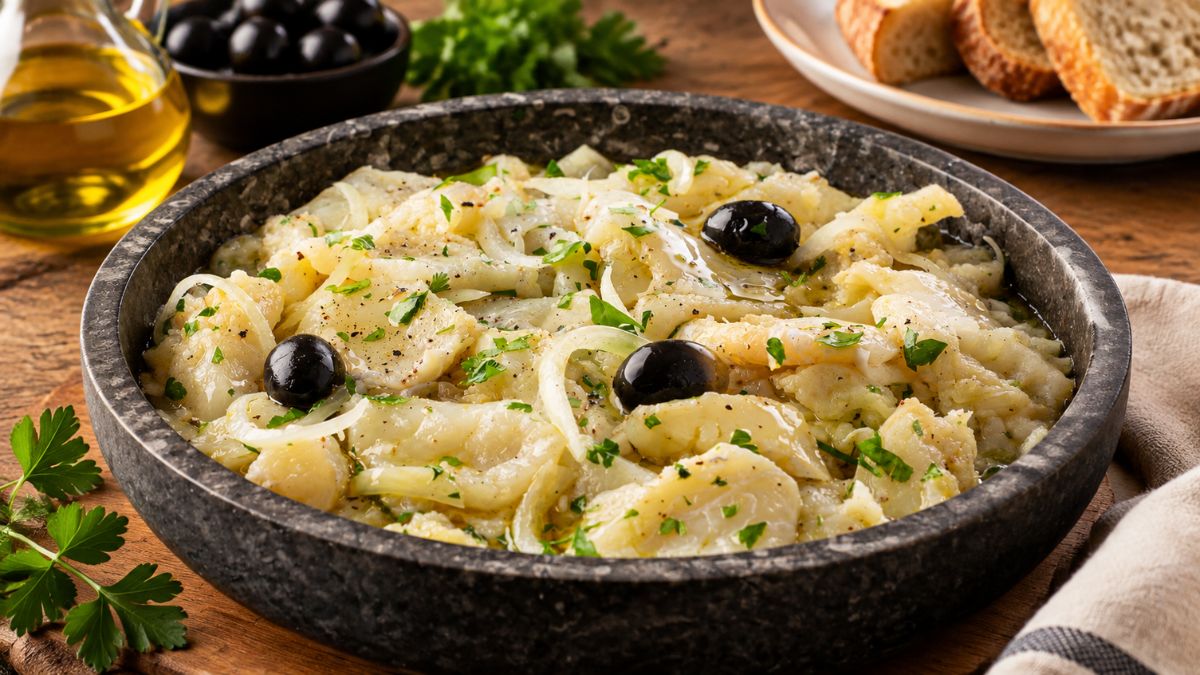 Uma tigela de pedra escura rústica está cheia de Punheta de Bacalhau, uma salada tradicional portuguesa feita com lascas de bacalhau demolhado, cebola fatiada fina e salsa fresca picada. O prato é regado generosamente com azeite e decorado com azeitonas pretas inteiras. Ao fundo, uma tigela de azeitonas pretas, um galheteiro de azeite e fatias de pão rústico completam a cena em uma mesa de madeira.