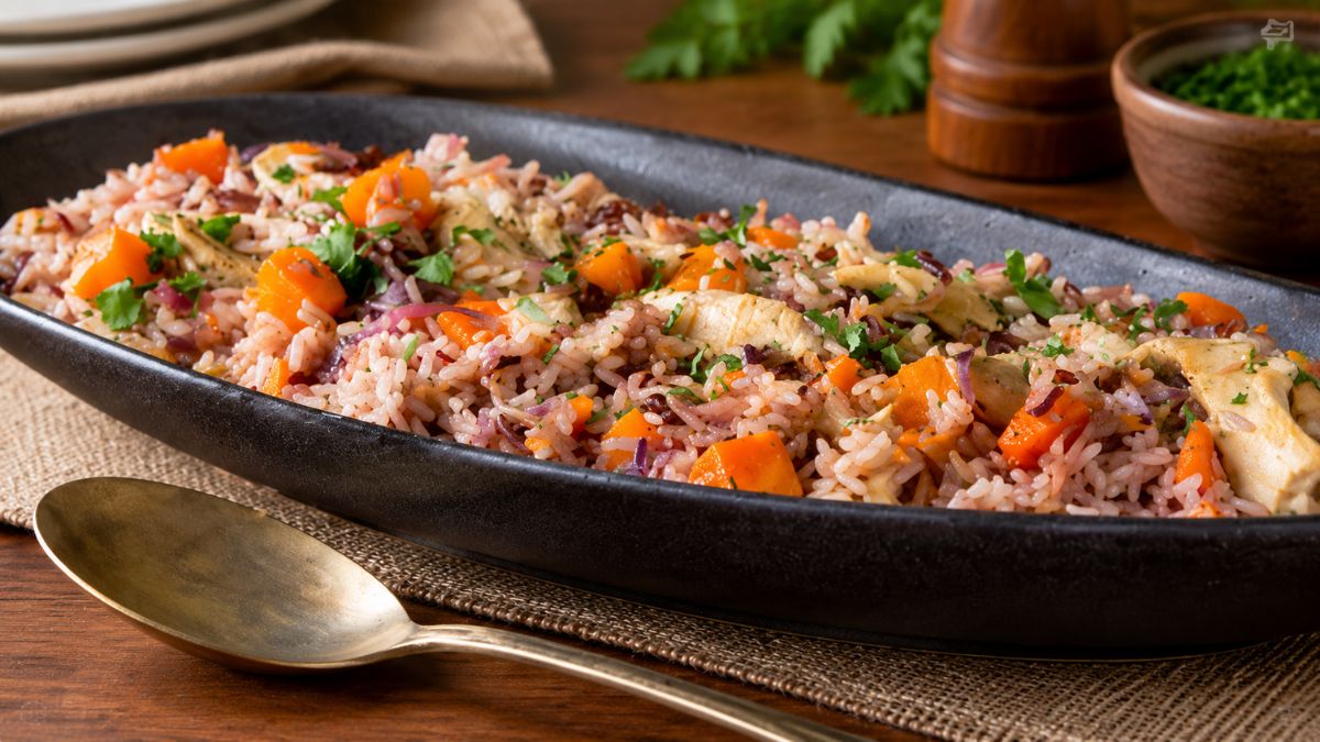 Uma travessa rústica preta servida sobre uma mesa de madeira, contendo arroz de frango solto misturado com cubos de abóbora assada, tiras de couve roxa refogada e decorado com salsa picada fresca. Há uma colher vintage dourada ao lado e ingredientes ao fundo.