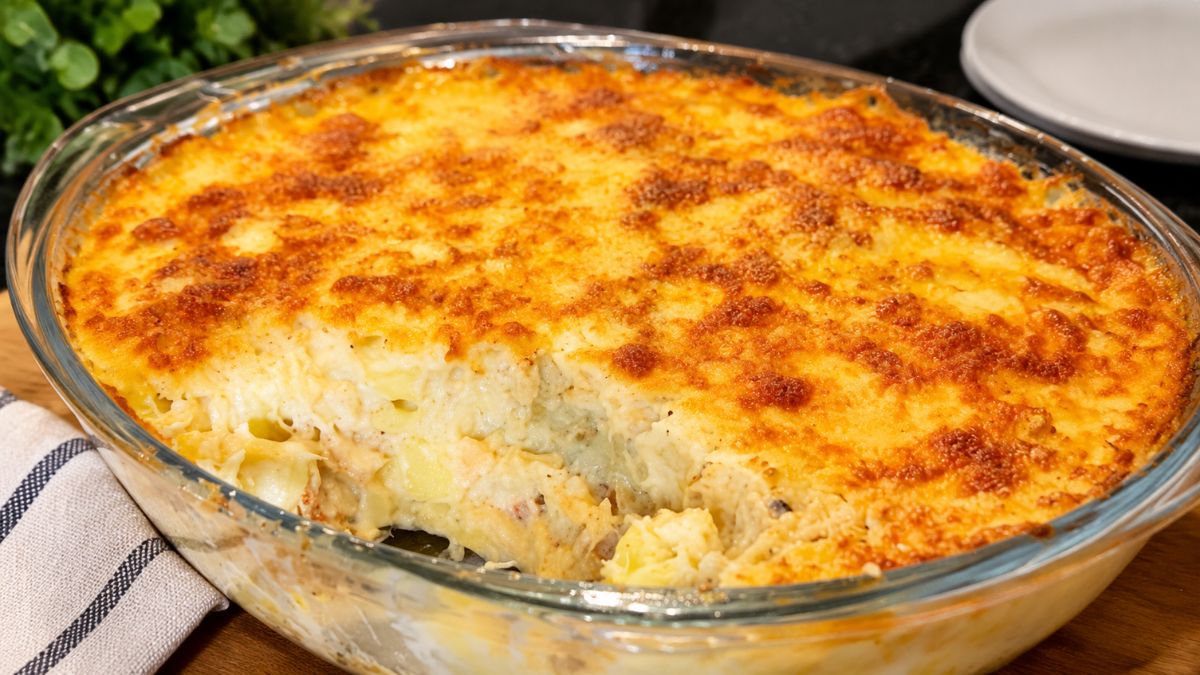 Fotografia em close-up mostra uma travessa de vidro transparente preenchida com bacalhau com natas. O gratinado de queijo dourado por cima está parcialmente partido, revelando o recheio branco, cremoso e texturizado com bacalhau e batatas lá dentro. A travessa repousa sobre uma mesa de madeira.